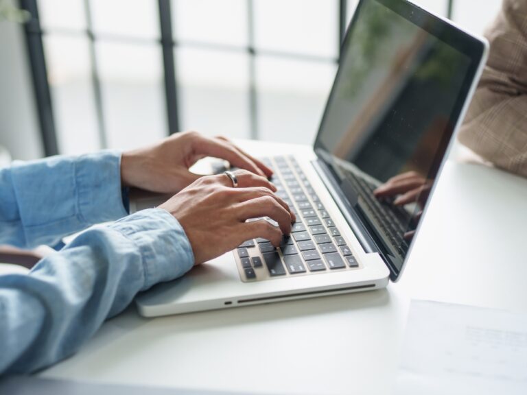 Person typing on a silver laptop at a white desk, wearing a light blue shirt.