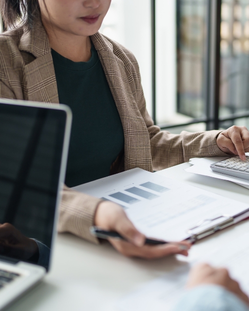 Professional desk scene: woman in a plaid blazer reviews documents with a calculator at a bright desk